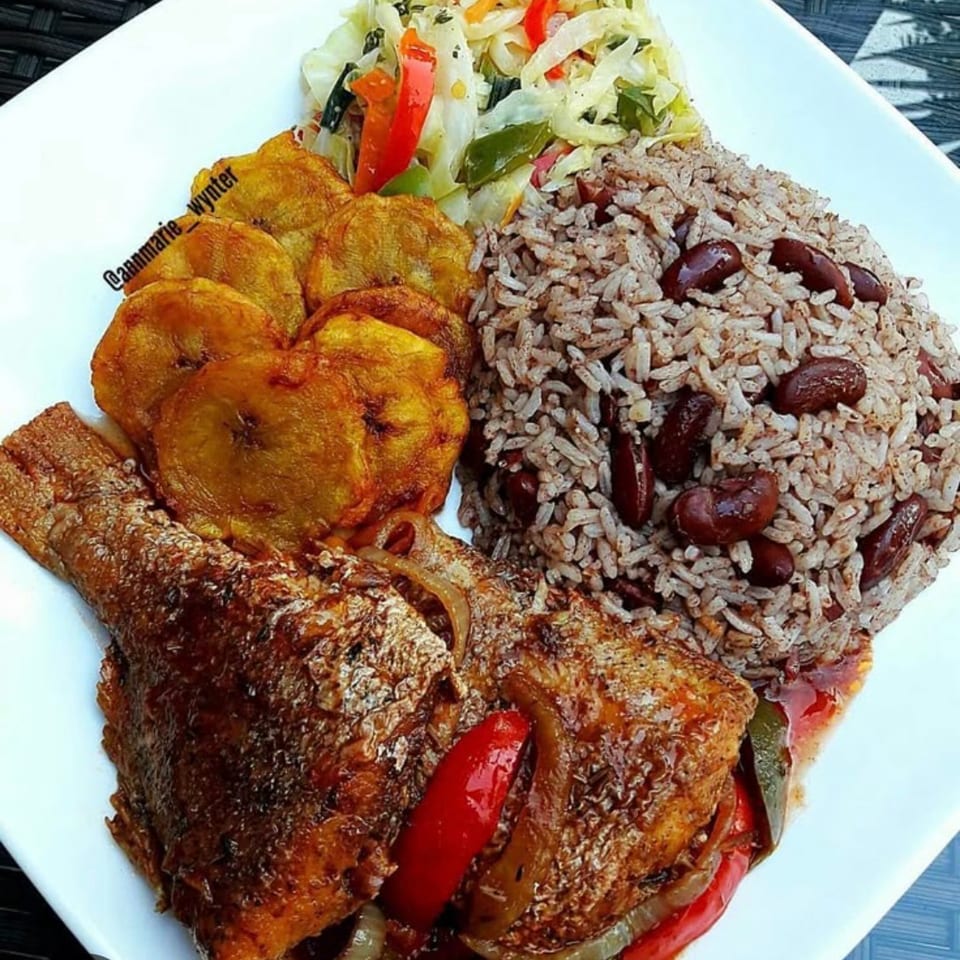 A plate of brown stew fish with rice and peas, fried plantains, and steamed cabbage.